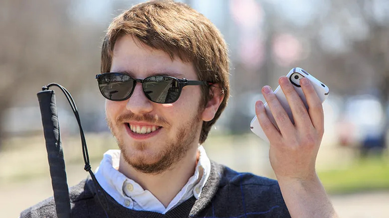 A smiling man wearing sunglasses and holding a cane aloft holds a phone in his other hand as though he is listening to the speaker.