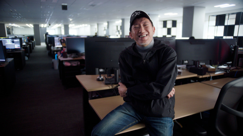 Shinji Mikami smiles while sitting on a desk at Unbound Inc., his studio.
