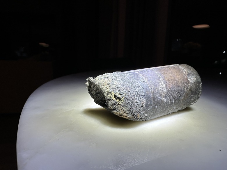 Photo of a stanchion from an ISS space dump. The object, a gray-ish cylinder with crumbled / burned edges, sits on a white table with black background.