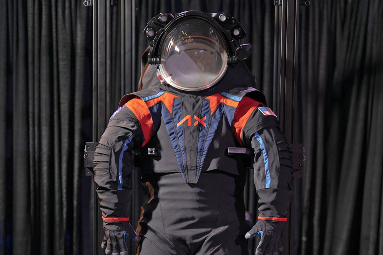 The Axiom Space AxEMU spacesuit for Artemis III Moon mission is presented against a black curtain. The suit is helmeted and mostly dark grey with blue and orange accents.