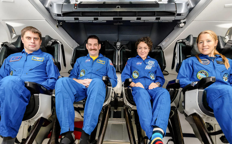 Four people wearing blue astronaut overalls sitting inside a spacecraft.