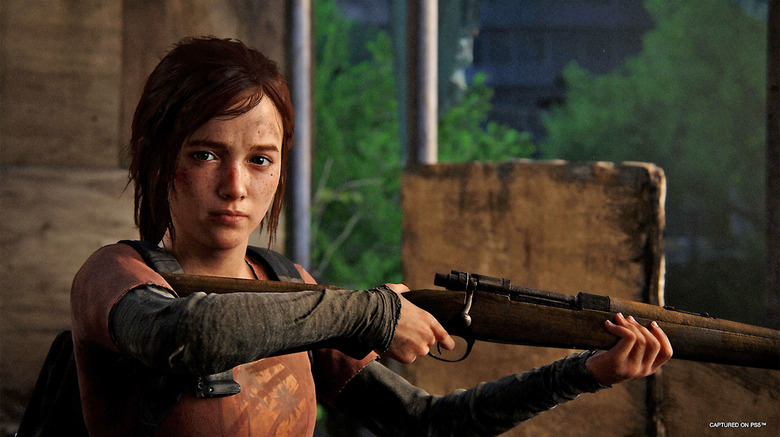 In this still captured via PS5, the Ellie character looks at the camera sideways while pointing a rifle out of the side of a wall opening.