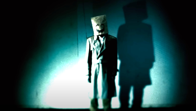 A still figure wearing a trench coat and a paper bag over their head with a smiling face drawn on it