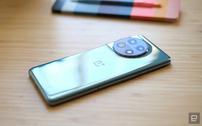 A green metallic OnePlus 11 5G smartphone sits face-down on a wooden table top with a pen and notepad in the background.