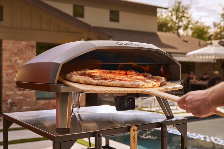 A person's hand using a pizza peel to remove a large cooked pizza from an outdoor pizza oven that's located on a patio.