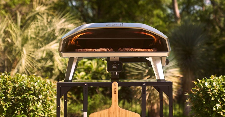 Ooni Koda 2 Max pizza oven sitting on a metal stand in an outdoor setting. Two pizza peels are hanging on the front of the stand and there's visible fire inside the pizza oven.