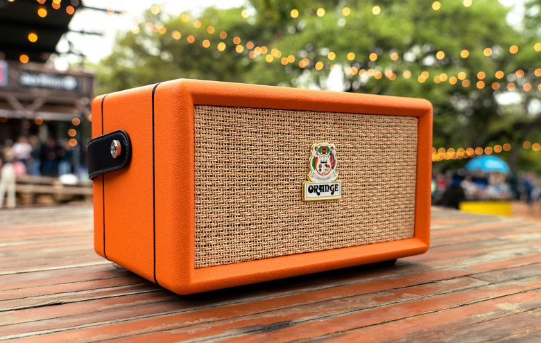 The Orange Box portable Bluetooth speaker sits on a wooden picnic table with string lights overhead, with a crowded outdoor event space and trees blurred in the background.