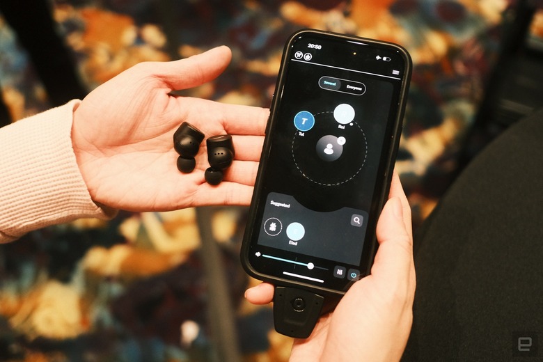 A pair of black earbuds on a person's palm, with their other hand holding up a phone with the OrCam Hear app on its screen.