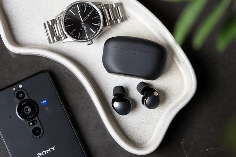Sony WF-1000XM5 earbuds in black pictured on a tray next to the charging case, a silver watch and a Sony smartphone