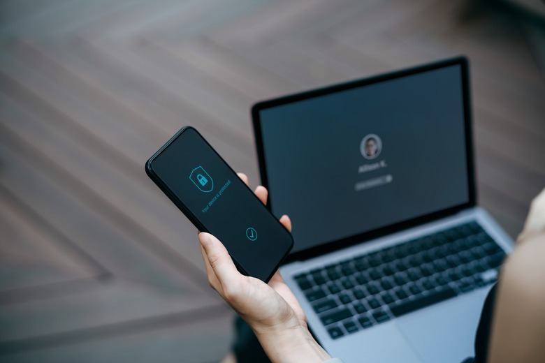 Close up, cropped shot of young woman sitting on the bench in urban office park working outdoors, logging in to her laptop and holding smartphone on hand with a security key lock icon on the screen. Privacy protection, internet and mobile security concept