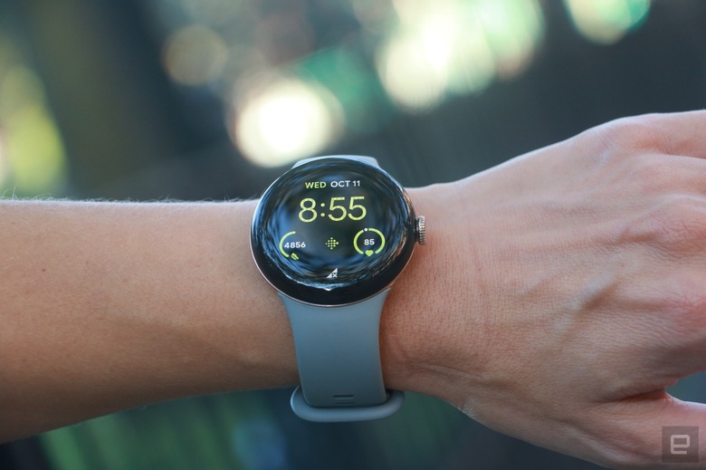 The Pixel Watch 2 on a person's wrist held in mid-air, with some blurred out greenery in the background.