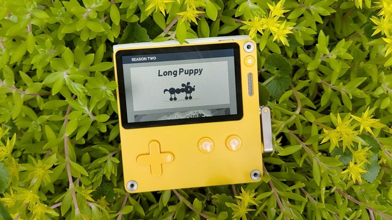 A Playdate handheld console is pictured lying in green and yellow foliage with the title card for the game Long Puppy displayed on its screen