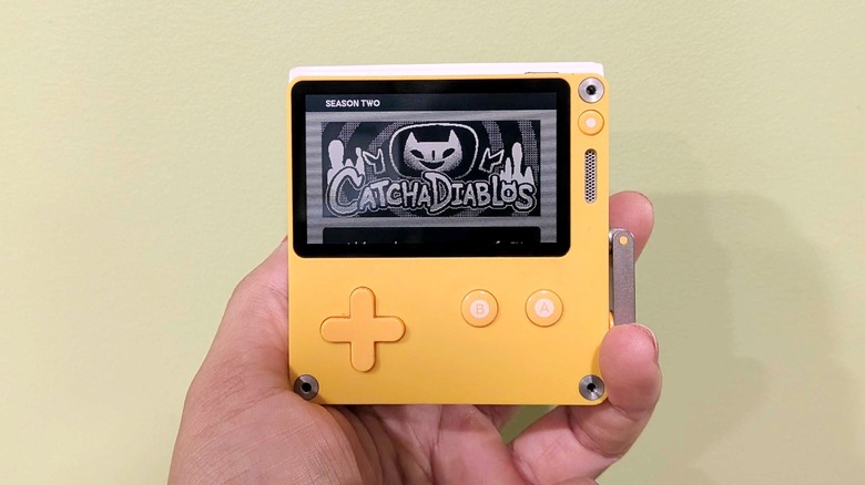 A Playdate console held in a person's hand up against a light green wall, with the screen displaying the title card for the game CatchaDiablos