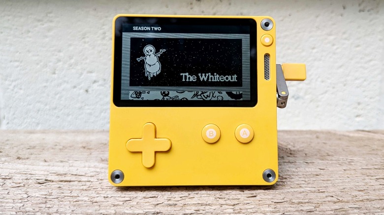 A yellow Playdate handheld console is pictured on a wooden surface displaying the title card for the Season 2 game, The Whiteout, which features a snowman with X'ed out eyes