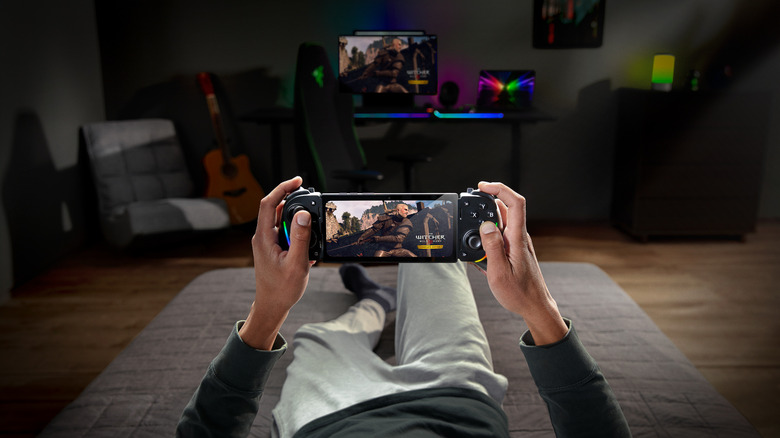 A person lying down on a bed, playing The Witcher on a mobile device. The host PC is seen blurred in the background.