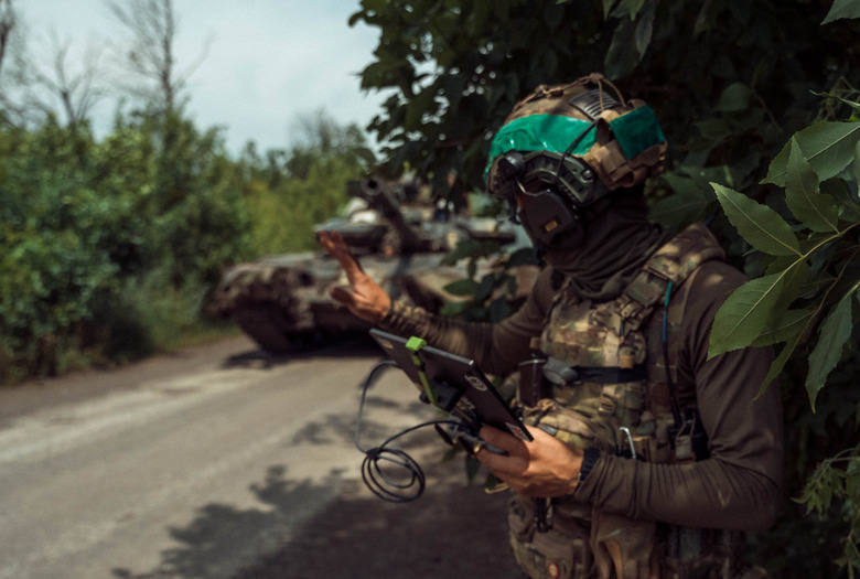 A Ukrainian soldier operating a drone. Forest, next to a street. The solider is holding a tablet with wires as a friendly tank sits behind.
