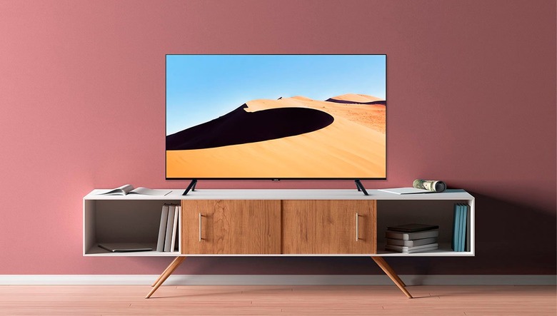 A press image of the Samsung TU690T 4K TV, displaying a TV on a white and brown cabinet against a light red wall. The image on the TV displays a desert dune.