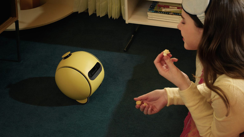 A young person, eating popcorn, looks at a yellow Ballie robot in the center of the frame.  