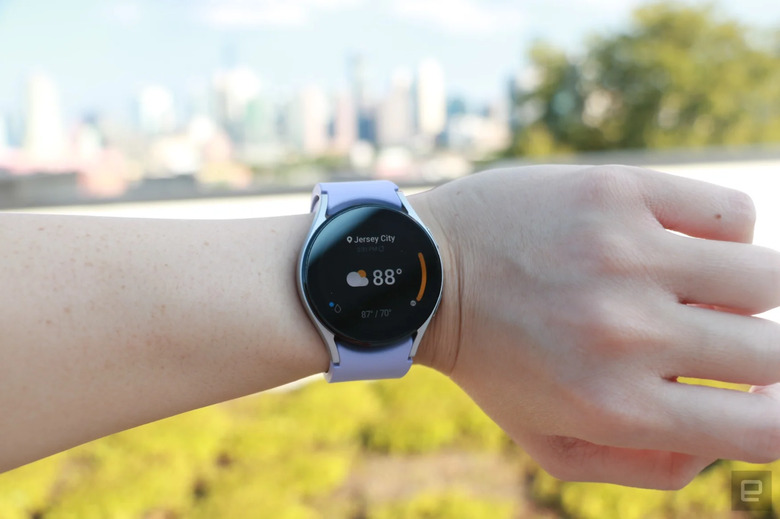 Photo of an arm wearing the Samsung Galaxy Watch 5 with lavender band against a background of trees, grass and buildings.