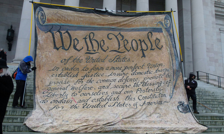 Protestors stand on the steps of a government building, holding a supersized banner recreation of the preamble to the US Constitution.