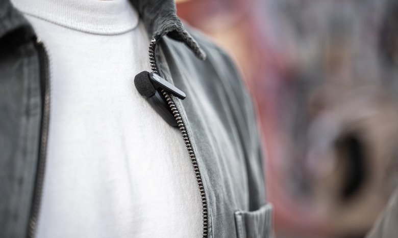 Product lifestyle image for the Shure MoveMic clip-on lavalier microphone. A person has the mic clipped to a gray-colored jacket with light material.