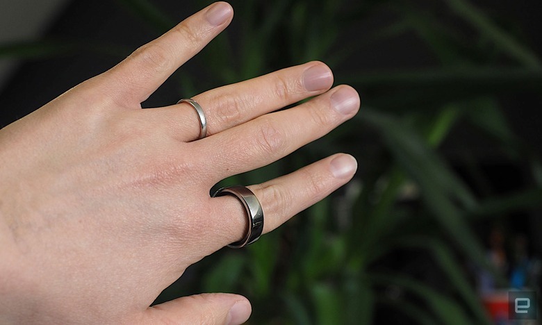 Image of a hand wearing an Oura Ring on the index finder in front of a dark green plant.