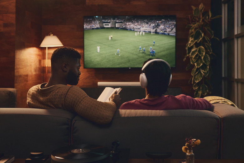 Two people (view from behind) sitting on a living-room couch with a TV on in the background. A soccer game is on in the background. One person is reading a book, while the other watches the game while wearing Sonos Ace headphones.