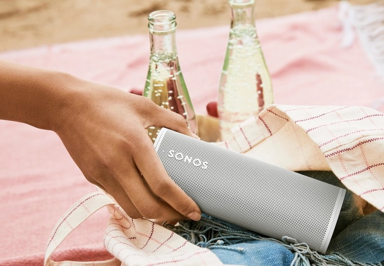 A closeup of a person's hand taking a Sonos Roam speaker out of a bag with bottles of a carbonated beverage behind it