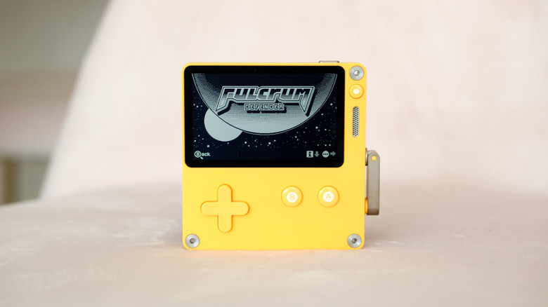 A Playdate sits on a pink seat, with the splash screen for Fulcrum Defender displayed