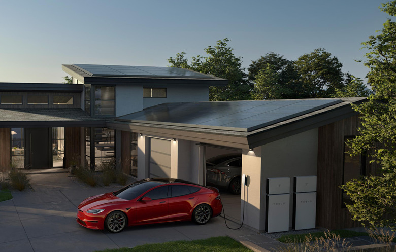 A house with solar panels on the roof. In front of the garage door is a red Tesla plugged into an outlet. White Powerwall units are visible on the outside wall of the garage.