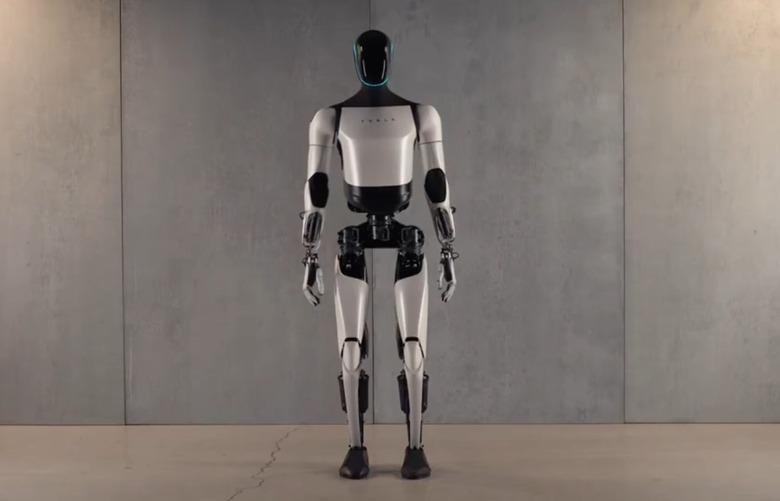 A humanoid robot with a Tesla logo on its chest placed against a concrete wall.