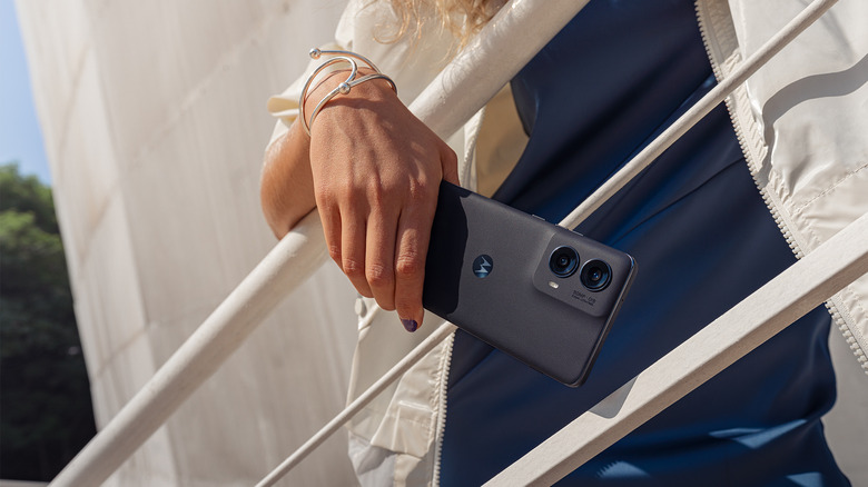 A Person in a white jacket leaning against a white stair railing (outdoors). The 2024 Moto Edge phone is in their hand.