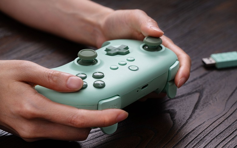 A person holding a lime-green wireless gamepad. A green USB dongle sits behind it on a dark wooden table.