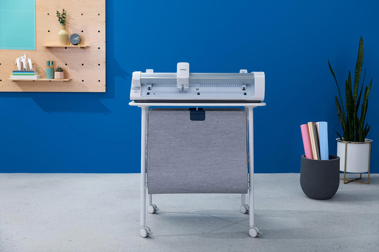 Marketing photo of the Cricut Venture, a consumer and small-business cutting machine for craft projects. It sits on a stand with a drape of fabric hanging below. It sits in a stylish room with a blue wall, gray floor and modern motif.