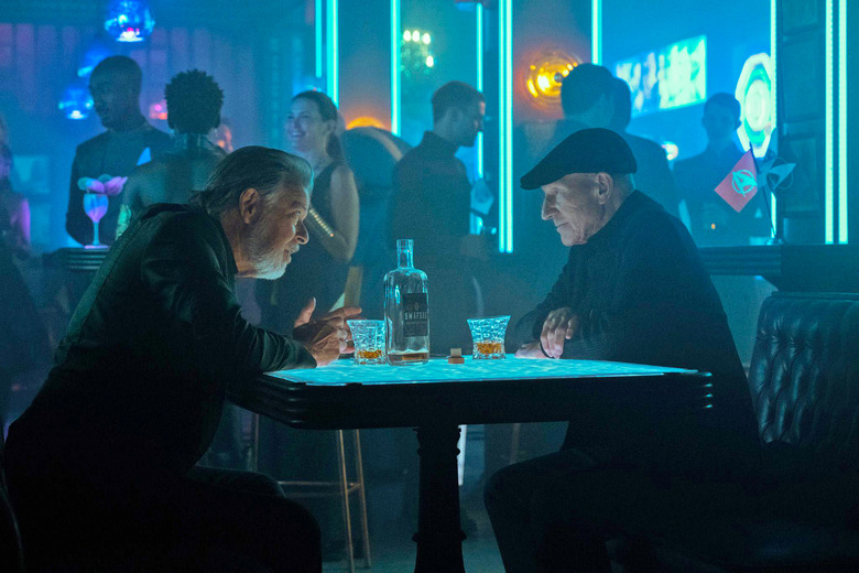 A still image from the TV series 'Star Trek: Picard Season 3' showing Picard and Riker sitting in a neon-lit bar at a table drinking liquor in tumbler glasses with the bottle on the table between them.