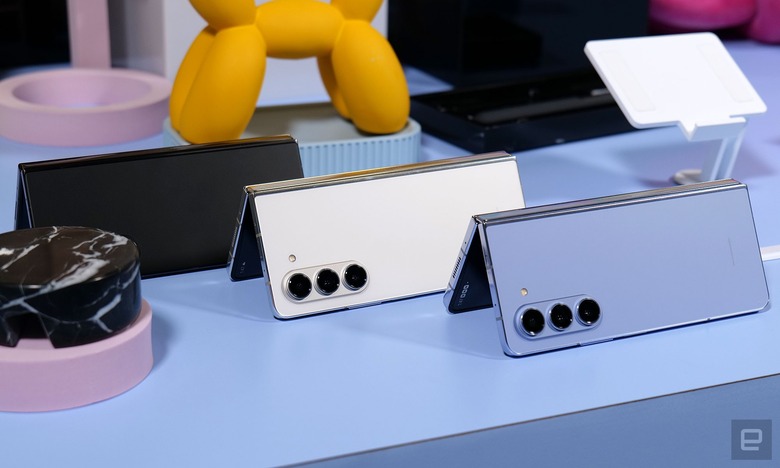 Three colorways of Samsung's Galaxy Z Fold 5 foldable smartphones are lined up on a blue table top with colorful knick·knacks in the background. 