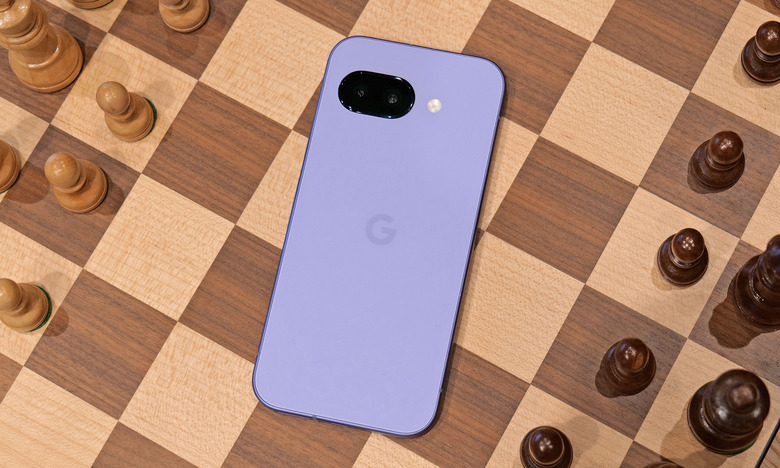 The purple Pixel 9a laying on the middle of a chess board, with black and white chess pieces on either side. The phone's screen is facing down.