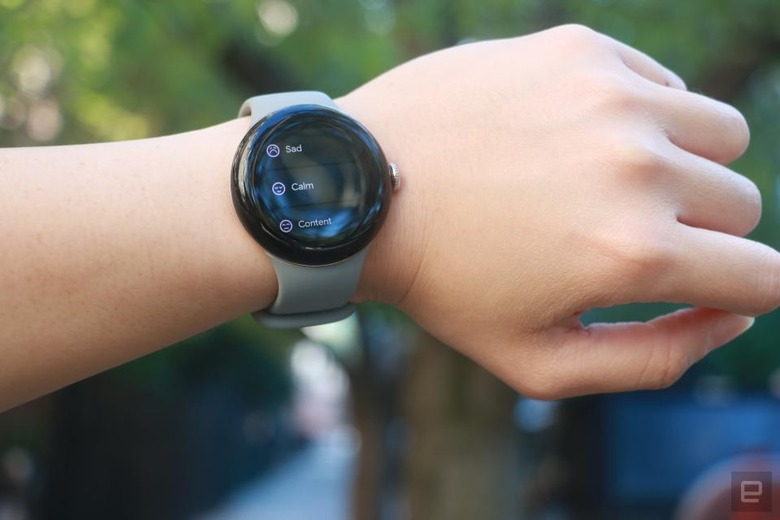 The Google Pixel Watch 2 on a person's left wrist, which is held up horizontally. The screen shows a list of feelings to log, including 