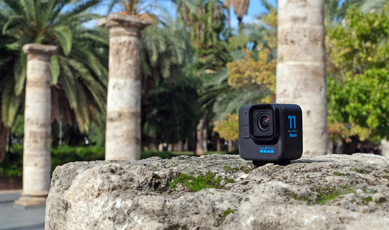 The GoPro Hero 11 Black Mini pictured on a rocky surface in a park with roman-style columns and palm trees in the background.