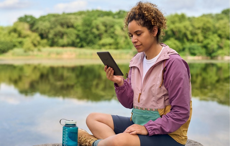 A woman sits on a rock near a lake reading from an Amazon Kindle