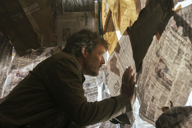 Joel (Pedro Pascal) peers through a gap in a window covered by newspaper in The Last of Us.       