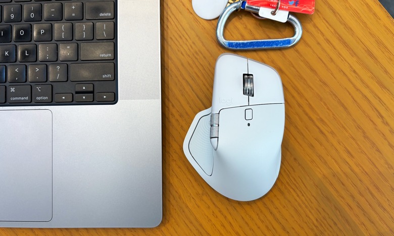 A white wireless mouse, the Logitech MX Master 4, rests on a brown wooden tabletop next to a blue set of keys and a silver laptop.