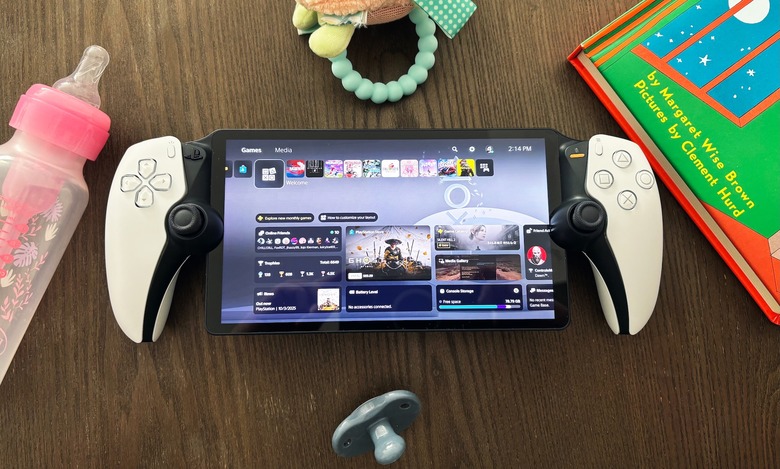 The PlayStation Portal gaming handheld sits on a brown wooden table surrounded by various parenting items, including a bottle, teething toy, pacifier and children's book.
