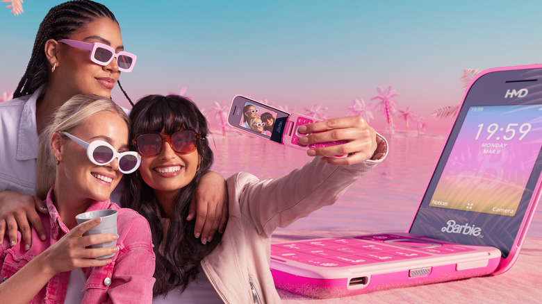 Three people take a selfie with the Barbie Phone. A large version of the phone sits on a Malibu beach behind them.
