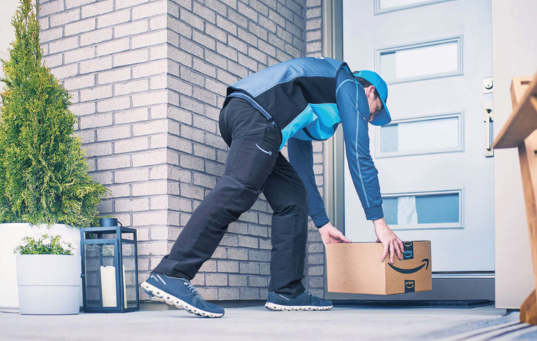 A person placing an Amazon-branded box on a welcome mat in front of a white door.