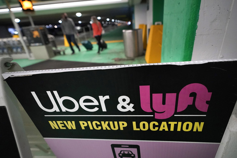 Passers-by pull luggage as they walk near a sign advising travelers of an Uber and Lyft pickup location at Logan International Airport, in Boston, Tuesday, Feb. 9, 2021. (AP Photo/Steven Senne)