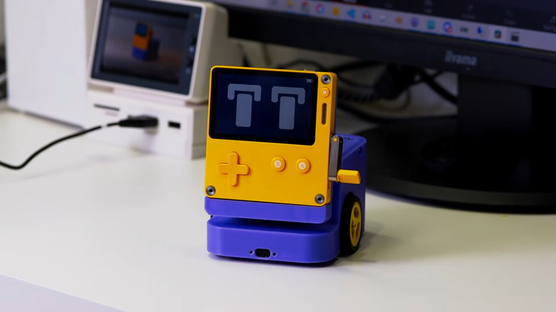 A Playdate handheld gaming console on a motorized dock with wheels. Its screen has cartoon eyes. It sits on a white desktop in front of a computer.