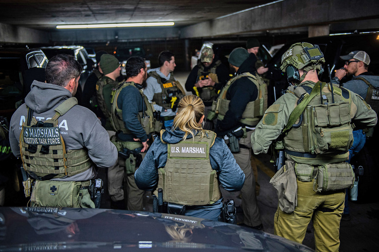 A group of US Marshals Service agents in Operation North Star II gather together in the dark depths of a parking garage with the hood of a car in the foreground.