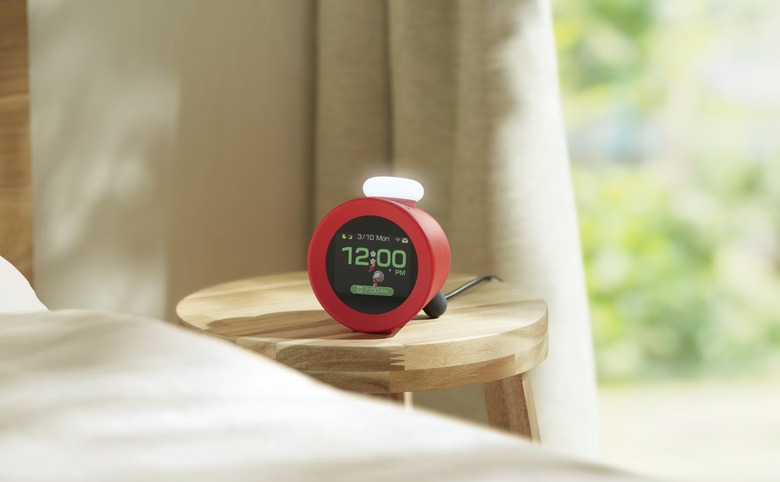 A Nintendo Alarmo clock is pictured on a bedside table in a sunlit room, displaying the time (12:00pm) and an animation of Olimar and a red Pikmin from the Pikmin franchise