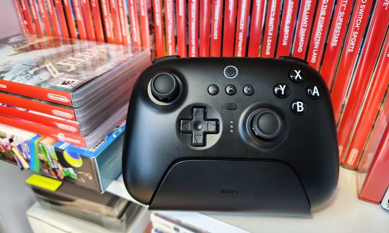 A black video game controller, the 8BitDo Ultimate Bluetooth Controller, rests on a white shelf in front of a row of several red Nintendo Switch video game cases.